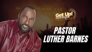 Pastor Luther Barnes smiles at camera wearing burgundy shirt on Get Up! Mornings with Erica Campbell show set with silhouetted audience and sun logo.