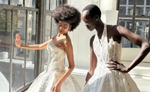 Two models wearing elaborate beaded white wedding gowns pose together in a sunlit high-rise space with floor-to-ceiling windows overlooking a city skyline.