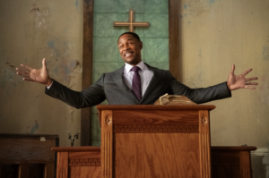 A man in a dark suit and burgundy tie stands at a wooden pulpit with arms outstretched, smiling broadly in front of a green-tiled wall with a copper cross.
