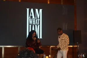 Two people sit on stage during an album playback event. A large screen behind them displays "I AM WHAT I AM" in illuminated white text. The woman on the left holds a microphone while the man on the right stands beside her.