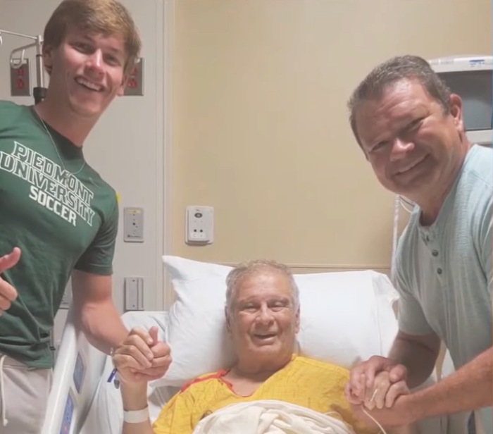 A grandpa in a hospital bed and his son and grandson holding his hands.