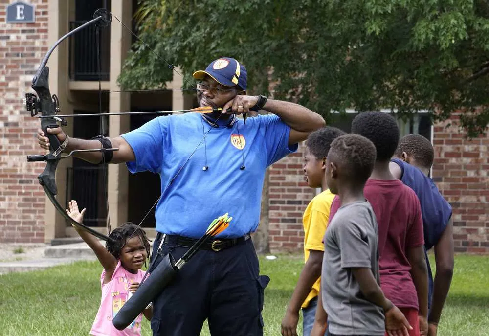 A police officer shooting a bow and arrow while children watch.