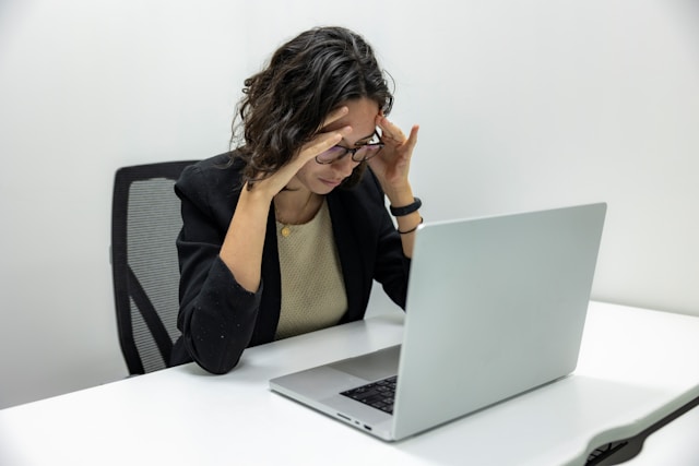 Frustrated woman with glasses holding her head while working on a laptop at a desk.
